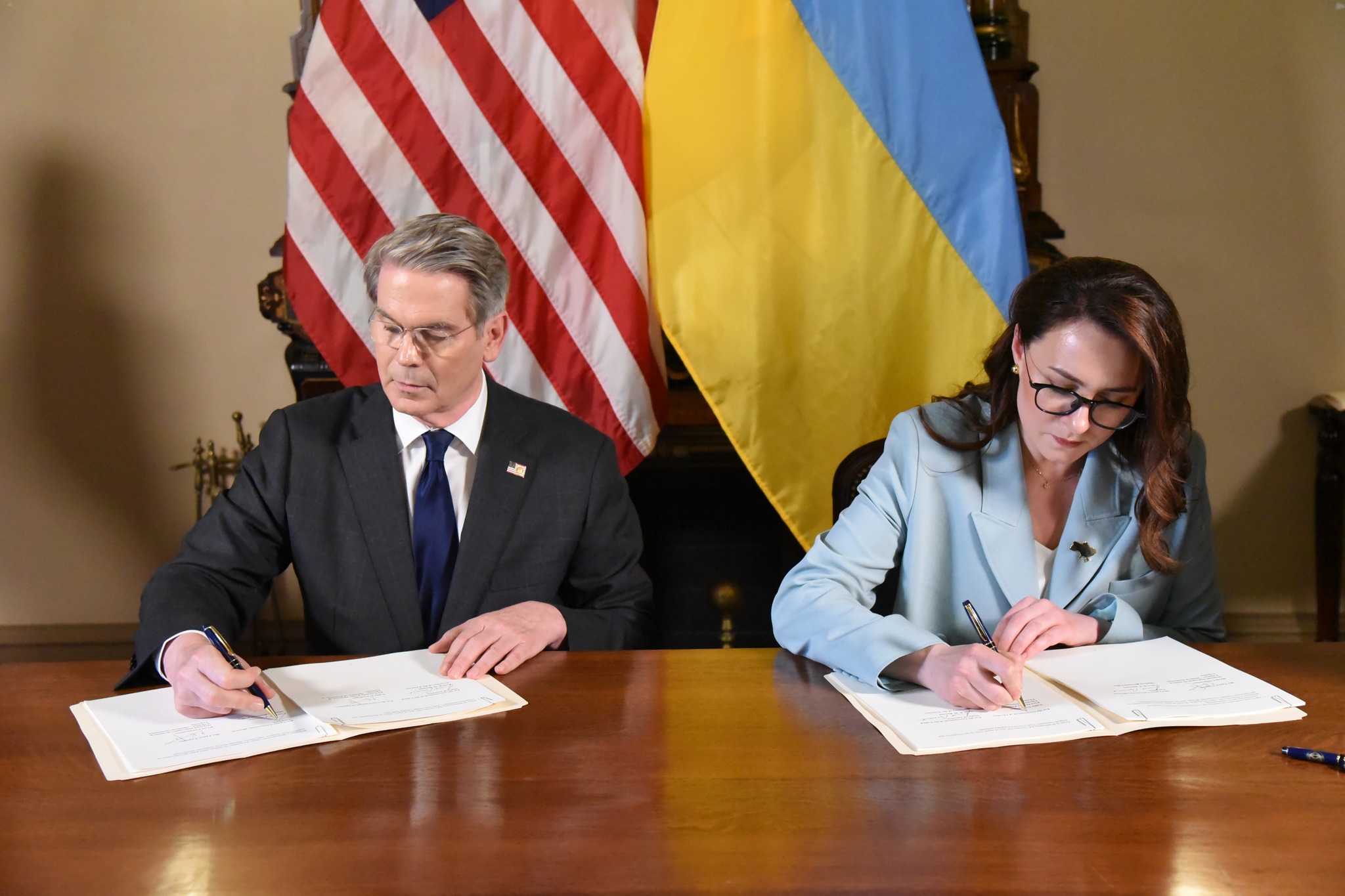 A male and female professional are both seated at a table signing documents in front of their respective flags (US and Ukraine).