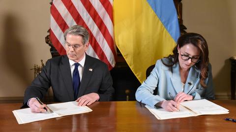 A male and female professional are both seated at a table signing documents in front of their respective flags (US and Ukraine).