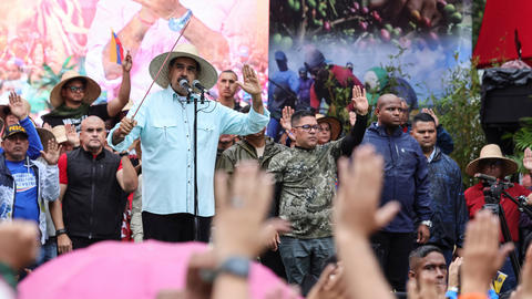 Venezuelan President Nicolas Maduro, wearing a straw hat and holding a stick, speaks at a microphone among an outdoor crowd with people raising up their left hands.