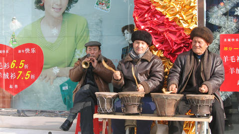 Three male Uyghur musicians sitting on sidewalk in front of a billboard, playing drums and horn