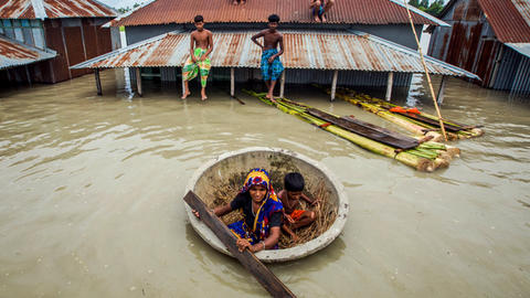 People sit on rooftops and use a makeshift boat in a flooded area with partially submerged buildings.