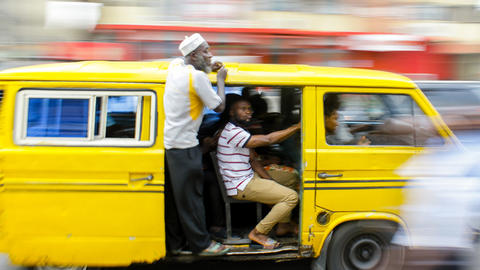 A bright yellow minibus (danfo) whizzes past, with the side van door open and two men hanging on.