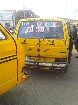 Rear view of a yellow minibus with a bumper sticker that says THE PRESENCE OF GOD stopped at a traffic stop with officers.