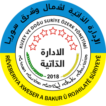 Circular emblem of the AANES shows a blue upper and yellow lower outer ring with Syrian words and seven red outlined stars inside.