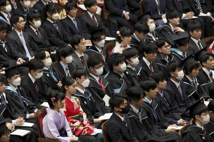 Dozens of Japanese college students, many of whom are wearing medical masks, are seated at a college graduation ceremony.