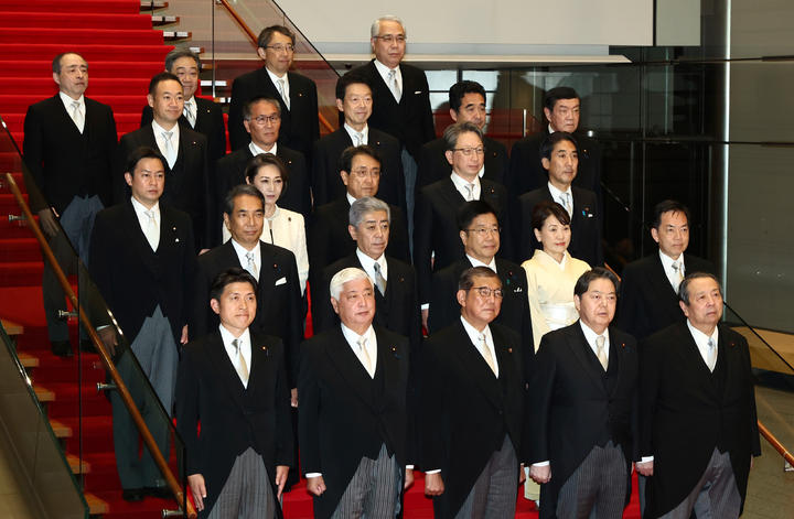 About twenty member of a Japanese cabinet—all dressed in suits, with only two women—stand on a red carpeted set of stairs.