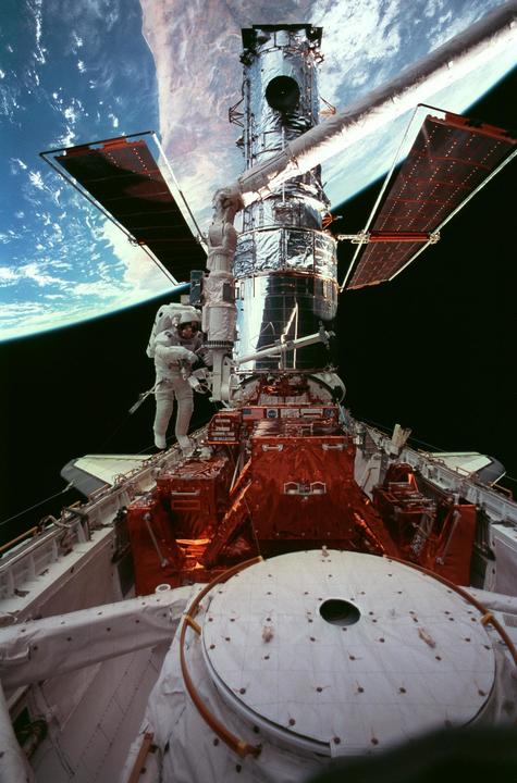 Astronauts working on the Hubble Space Telescope in space, with Earth in the background.