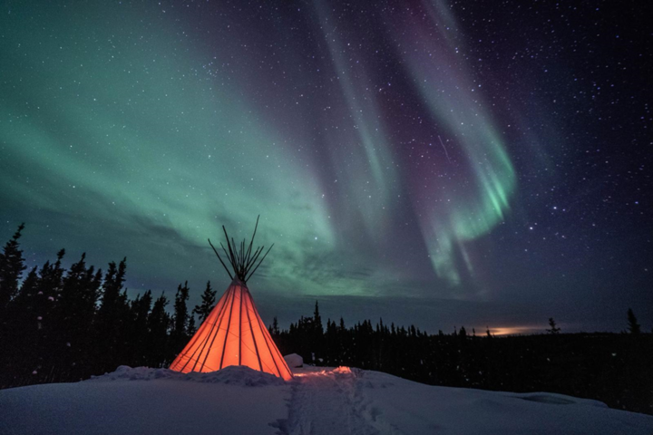 Glowing tipi in a snowy forest clearing with aurora borealis lighting up the sky.