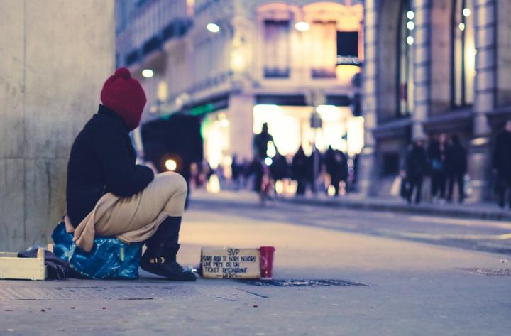 Person wearing winter weather clothing, sitting at a street corner at night with a cup and sign in front of them.