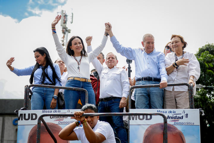 Two Venezuelan opposition leaders stand on a platform with others behind them and are holding hands above their heads and waving to a crowd.