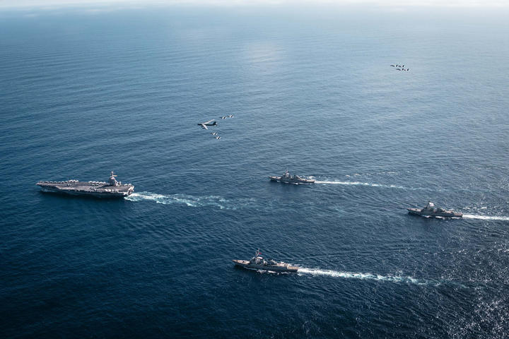 Aerial view of four US Navy ships, and a handful of airplanes, over the ocean.