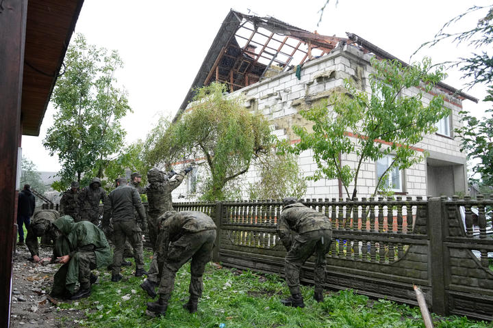 People in army fatigues are bending down to clean up debris in a yard adjacent to a house where the roof is decimated. 