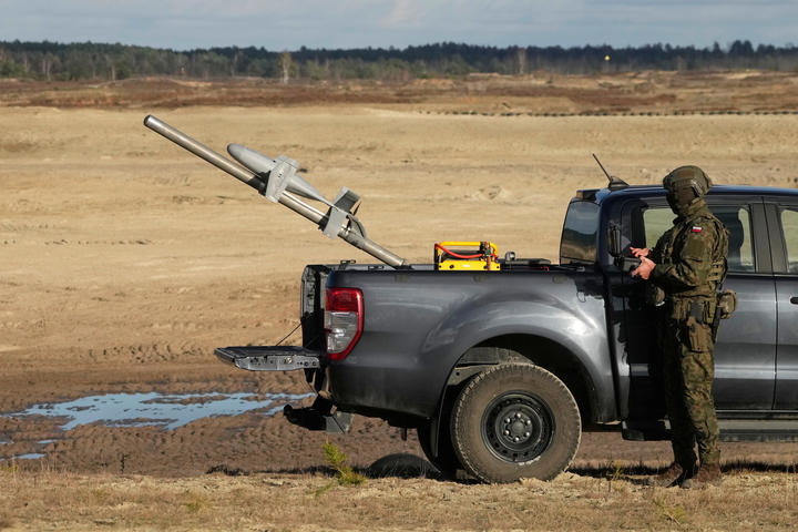 In a large dirt-packed area sits a gray truck with a drone in the bed, being operated by a person in army fatigues standing next to the truck. 