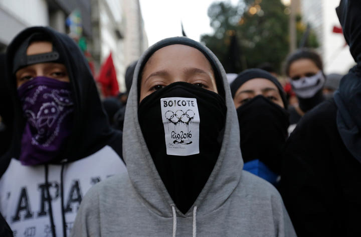 Several teenagers, wearing hoodies and face masks/bandanas, march in the street. The boy in the front is wearing a mask that reads in Portugues "Boycott Rio 2016." 