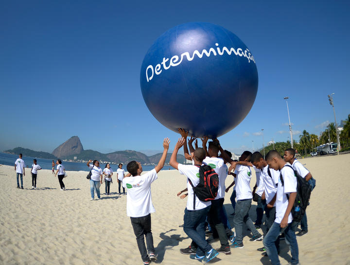 Several children are holding up a large blue balloon at the beach. 