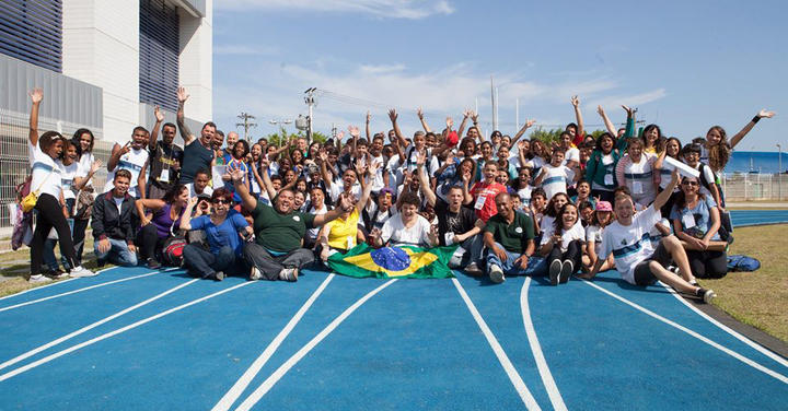 Several dozen people pose for a group photo, cheering and raising their arms, on a blue track located at a beach. 