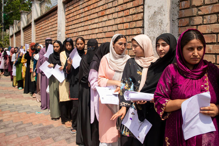Women wearing saris and head scarves stand in a long line next to a building while holding papers. 