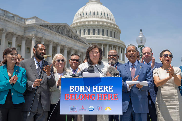 Members of Congress stand at a podium with a sign that says "BORN HERE BELONG HERE" in front of the US Capitol building. 