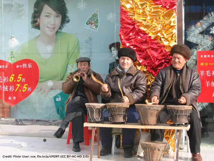 Three male Uyghur musicians sitting on sidewalk in front of a billboard, playing drums and horn