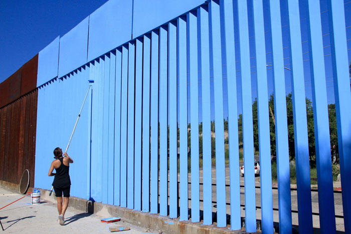 Image of an artist painting the porous border fence blue to match the sky
