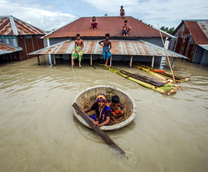 People sit on rooftops and use a makeshift boat in a flooded area with partially submerged buildings.