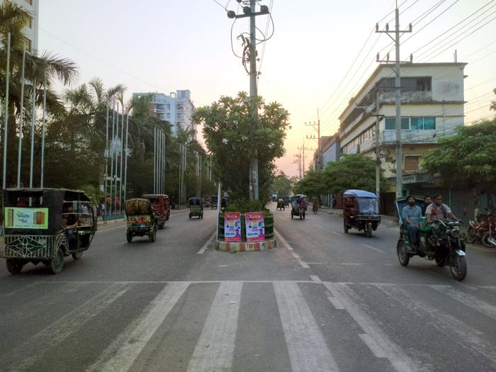 A city street at dusk with motorized rickshaws and a few pedestrians on either side. Buildings and trees line the road. A utility pole and a small intersection with some posters are in the center.