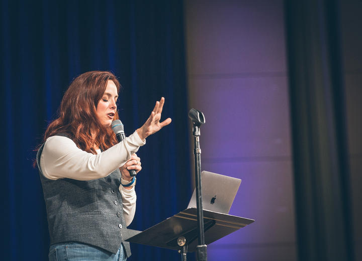Sarah Bessey praying while holding a microphone on stage.