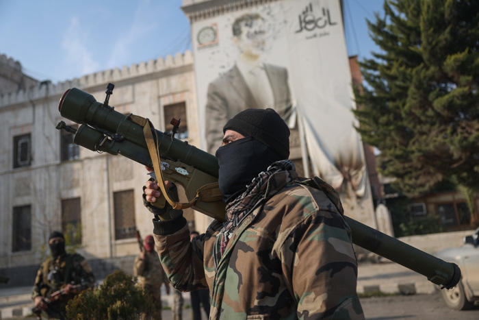 Profile photo of Syrian man holding weapon in front of a white building with a mural.
