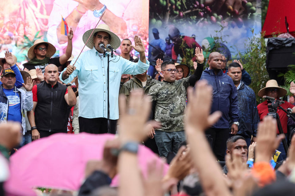 Venezuelan President Nicolas Maduro, wearing a straw hat and holding a stick, speaks at a microphone among an outdoor crowd with people raising up their left hands.